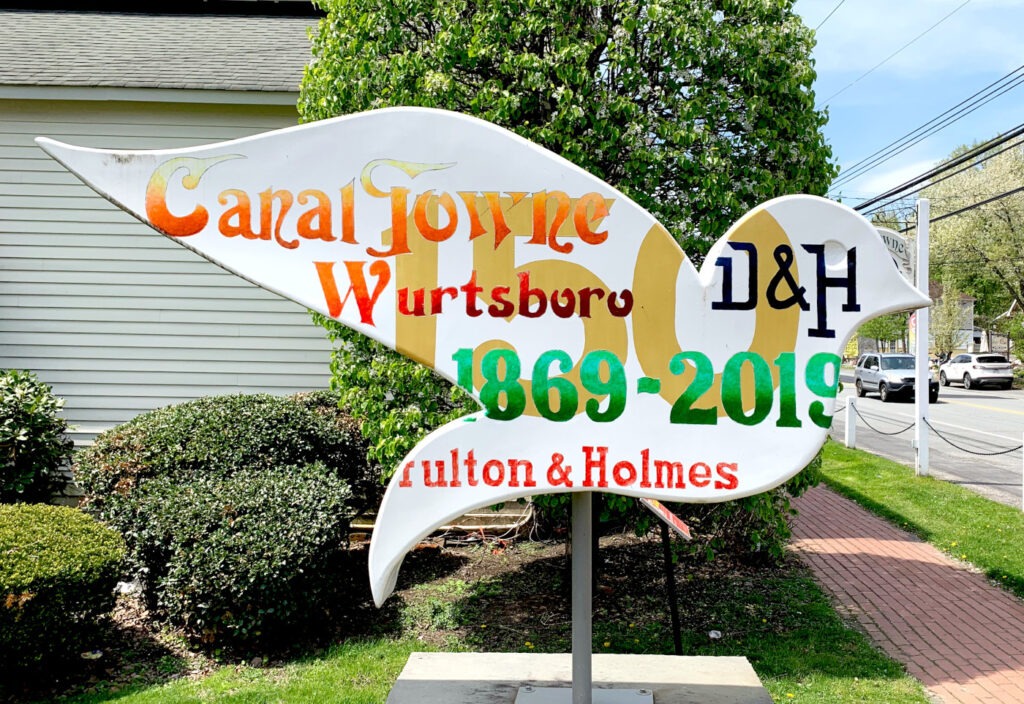 A dove-shaped sign, part of the Sullivan County Dove Trail, features the words "Canal Towne Wurtsboro" in orange and red, and "D&H 1869-2019" in green, with "Fulton & Holmes" below. The sign is set against a backdrop of green bushes and trees, with a road and cars visible in the background under a bright sky.