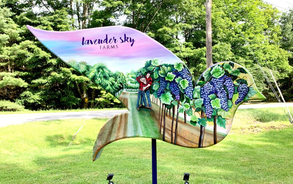 The reverse of the dove sculpture depicts a vineyard with a couple in flannel shirts embracing along a path between rows of grapevines. Clusters of purple grapes hang in the foreground, and the sky is softly painted in lavender and blue. “Lavender Sky Farms” appears on the upper wing.