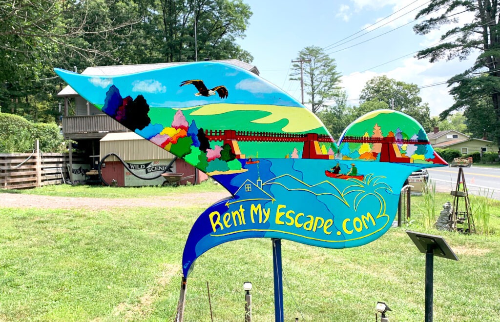 The opposite side of the same dove sculpture, painted with a vivid riverside scene. A couple in a red canoe paddles down the river beneath a red bridge surrounded by colorful trees. A stylized blue house and yellow lettering spelling “RentMyEscape.com” are featured in the lower portion. An eagle soars above rolling green hills and a bright blue sky.