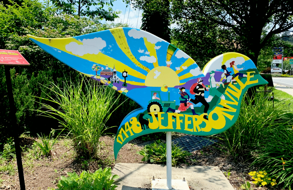 A brightly painted dove-shaped sculpture in Jeffersonville, NY, featuring colorful illustrations of community life. The artwork shows a sunrise with musical notes, children walking, a firefighter, a tractor, and local landmarks like a church and a clock tower. The word "JEFFERSONVILLE" is painted in bold yellow and green letters across the body, with "1748" on the dove’s tail. The sculpture is surrounded by greenery and flowers, with a red informational sign on the left and a road with traffic signs in the background.