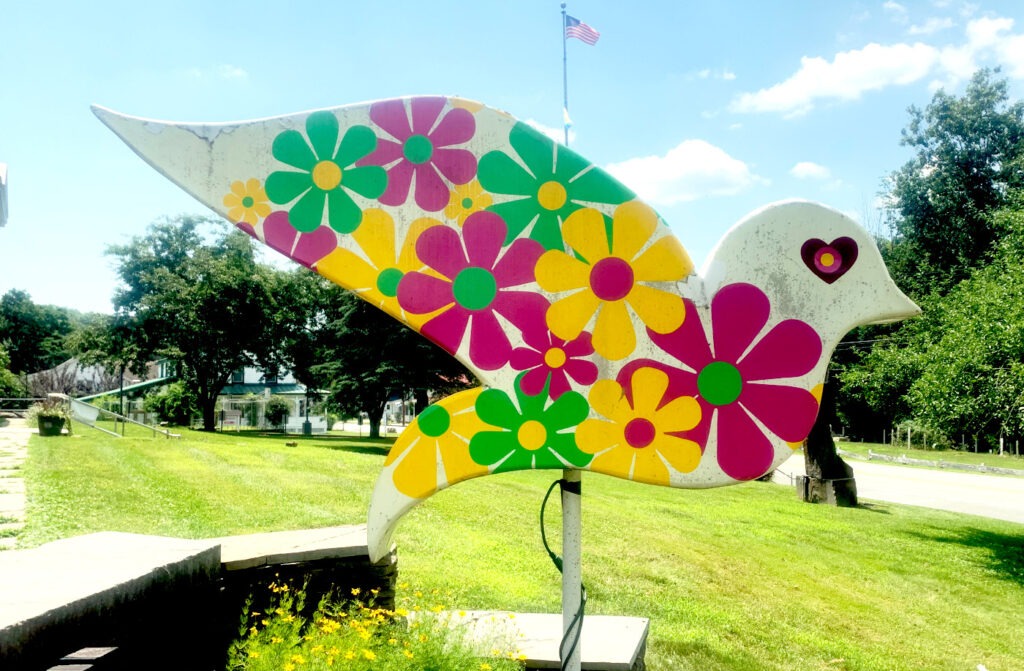 A white dove sculpture painted with a vibrant retro floral design featuring large daisy-style flowers in bright magenta, green, and yellow. The dove has a pink heart-shaped eye and stands on a grassy area with trees and buildings visible in the background.