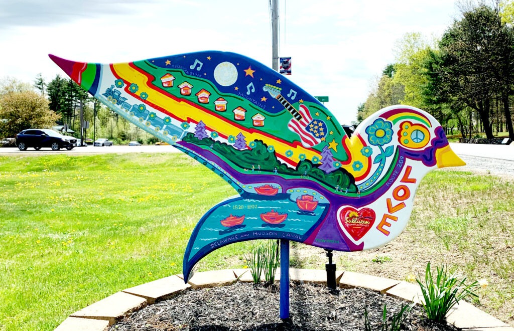 Mamakating dove sculpture displaying historical timeline elements including 'DELAWARE AND HUDSON CANAL 1828-1897' text, American flag imagery, houses representing local settlements, musical notes, stars, and nature scenes. The sculpture features bright rainbow colors, peace symbols, and floral designs with 'LOVE' text visible on the tail section."