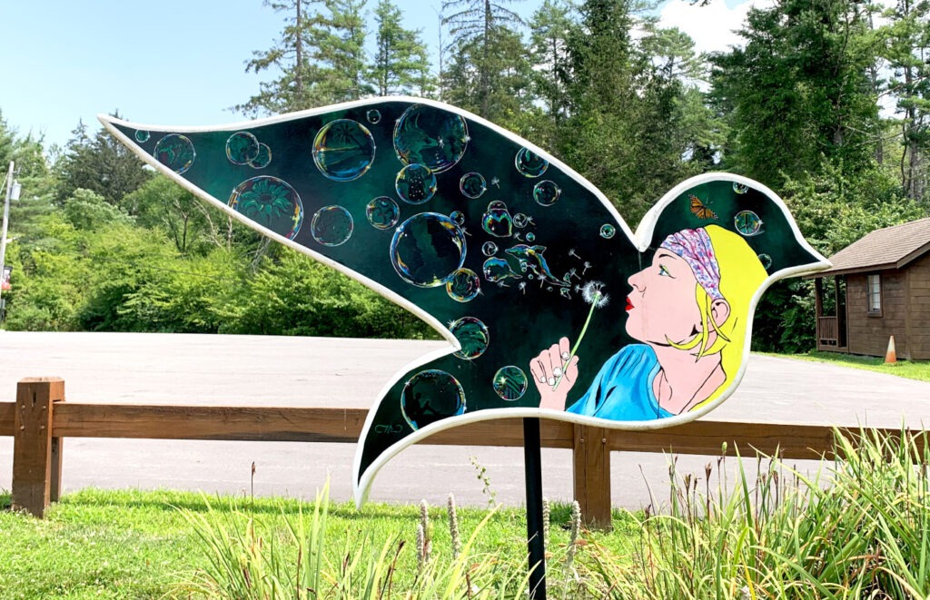 A dove-shaped sculpture in Circle Park behind Town Hall, painted dark green. It shows a blonde woman in a blue top blowing on a dandelion. The seeds drift upward and become translucent soap bubbles, each containing tiny scenic landscapes. A monarch butterfly flutters near her head. The sculpture is mounted in a grassy area in front of a wooden railing, with trees and a brown municipal building behind it.

