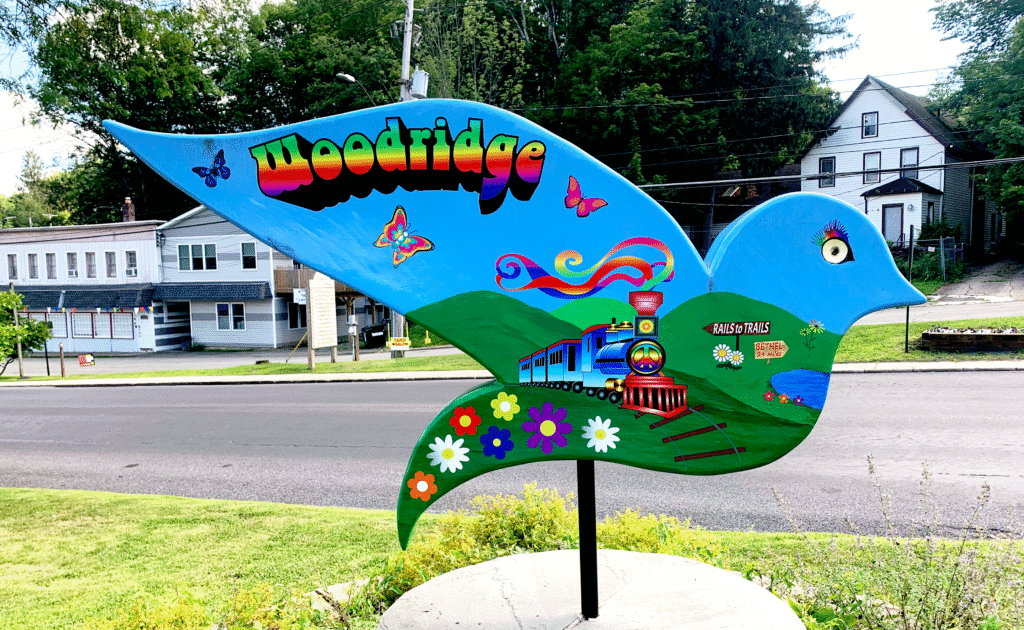 Reverse side of the Woodridge dove sculpture from the Sullivan County Dove Trail. The sculpture features a bright blue dove with colorful details, including a train with a rainbow steam trail and peace symbol, green hills, butterflies, and flowers. A sign on the hillside reads “Rails to Trails” and “Bethel 2.4 miles.” The word “Woodridge” is prominently displayed in bold, rainbow-gradient lettering across the wing. The background includes a small-town street with houses, storefronts, and a grassy lawn in the foreground.
