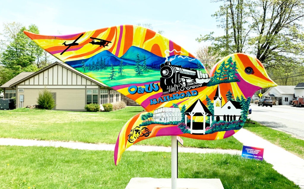 The reverse side of the Wurtsboro dove sculpture features a steam train labeled “O&W Railroad,” scenic mountains, vintage airplanes, a gazebo, a church with a bell tower, and a yellow motorcycle, highlighting the town’s transportation history and community landmarks.