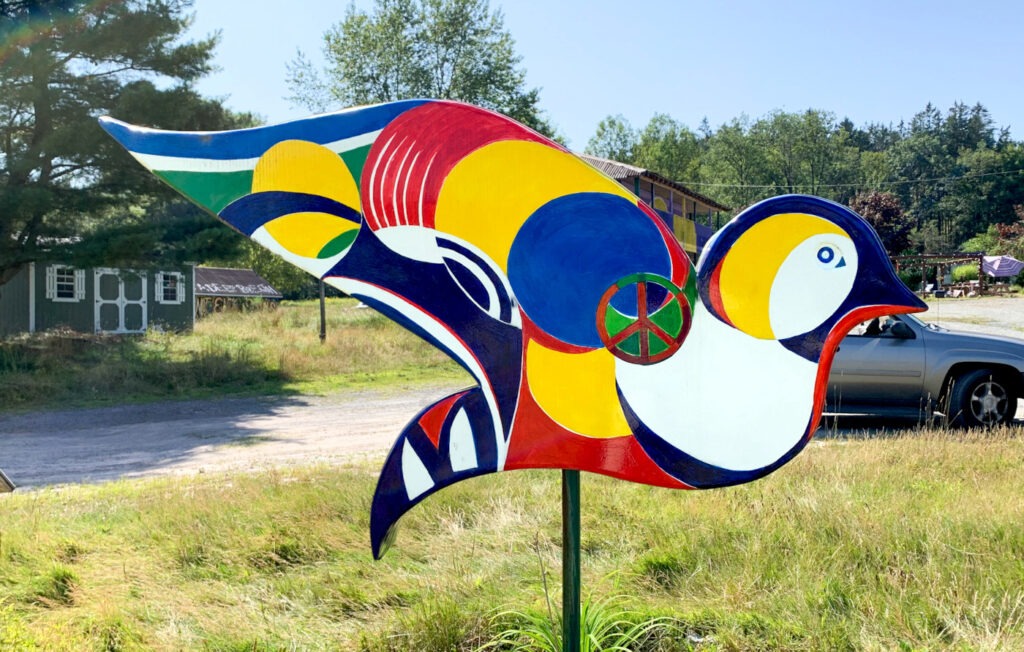 The dove sculpture displays flowing abstract shapes in bold swaths of yellow, blue, red, white, and green, centering around a peace symbol near the wing. The dove stands in a field with mixed rural structures and trees beyond.

