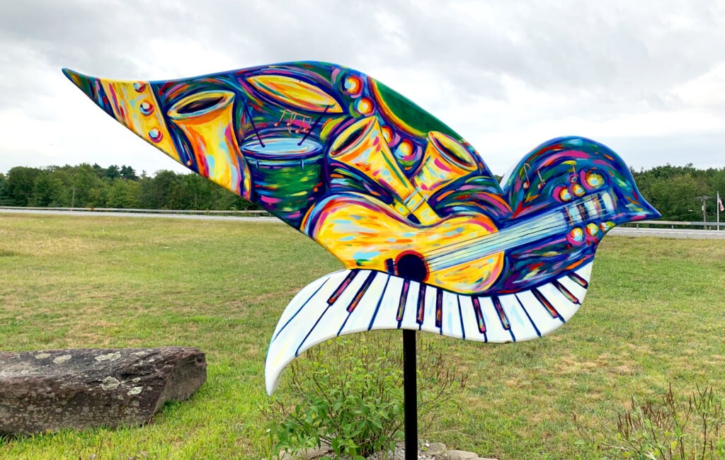 A dove sculpture, showing the continuation of the colorful musical instrument motif with drums, horns, and swirling artistic patterns in bright blues, yellows, oranges, and greens. The piano key pattern is visible on the tail section. The sculpture stands in an open grassy field with wooded areas and a road visible in the distance.
